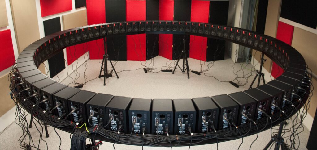 <em>Disposición circular de altavoces en sala tratada acústicamente, utilizada para reproducción inmersiva de campos sonoros y medidas experimentales de audio espacial.</em>